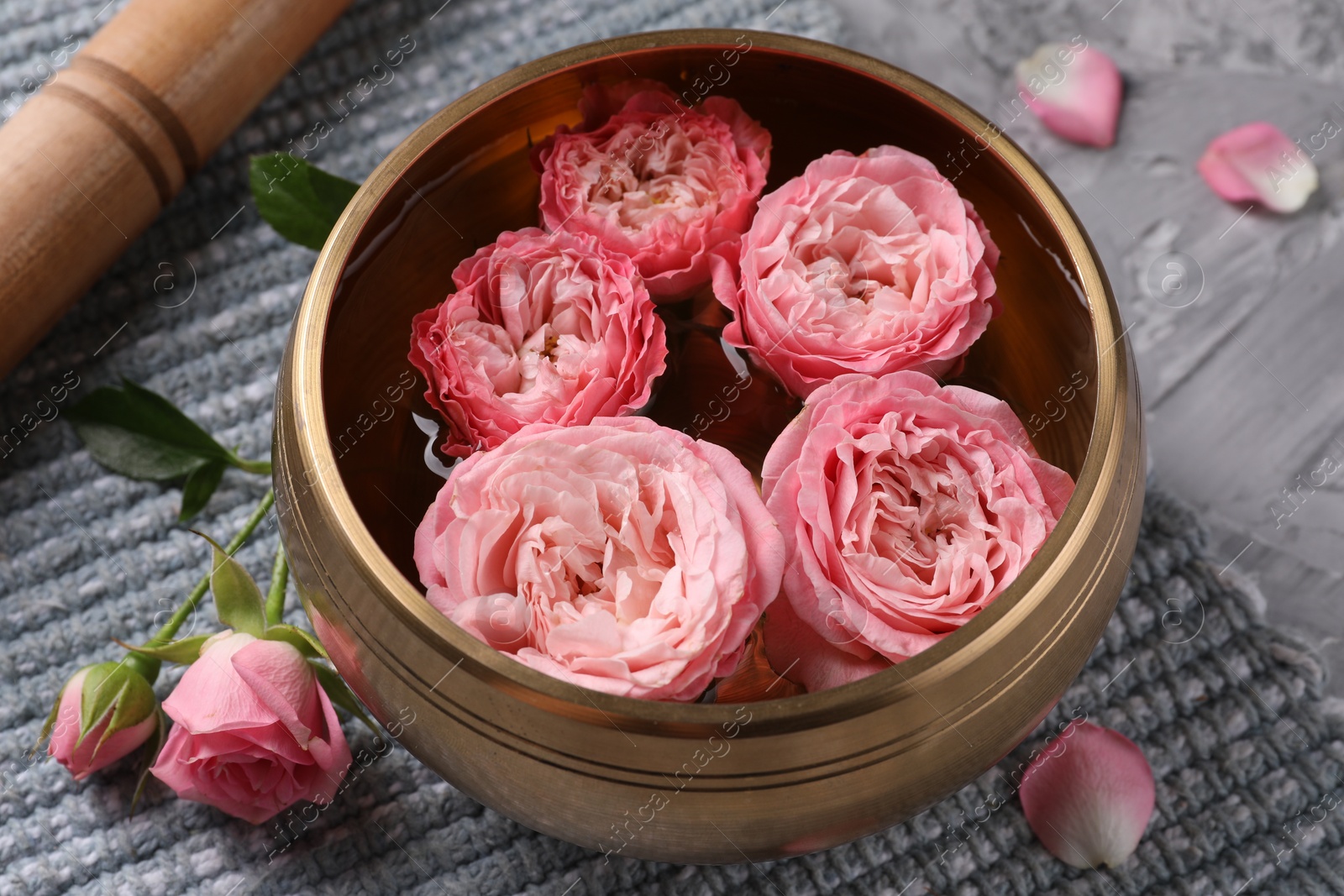 Photo of Tibetan singing bowl with water, beautiful roses and mallet on grey textured table, closeup