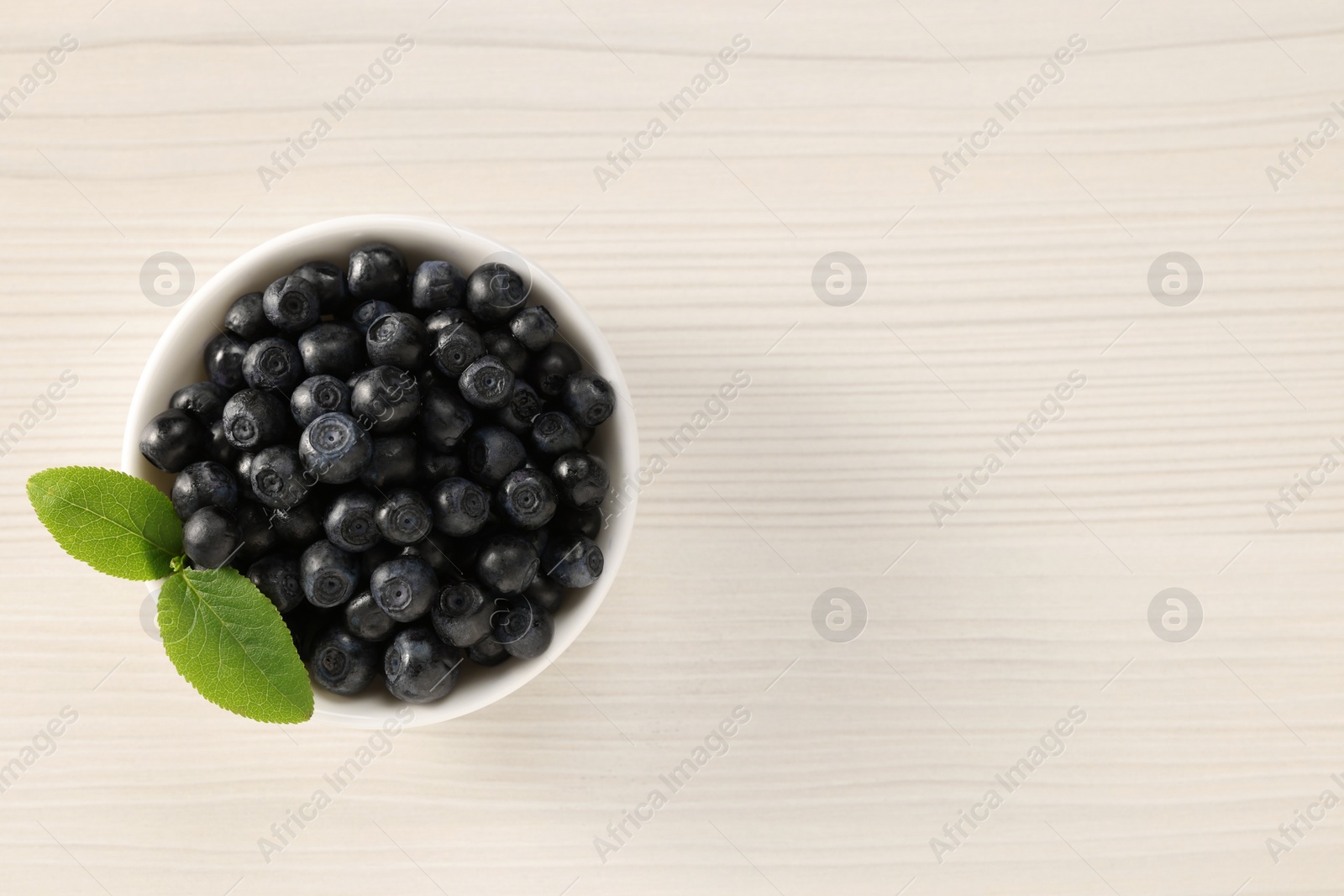 Photo of Tasty fresh bilberries with leaves in bowl on light wooden table, top view. Space for text