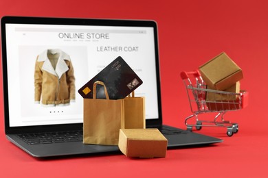 Photo of Online store. Laptop, small purchases, credit card and mini shopping cart on red background, selective focus
