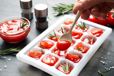 Photo of Woman pouring sauce into ice cube tray with tomatoes and rosemary at grey table, closeup