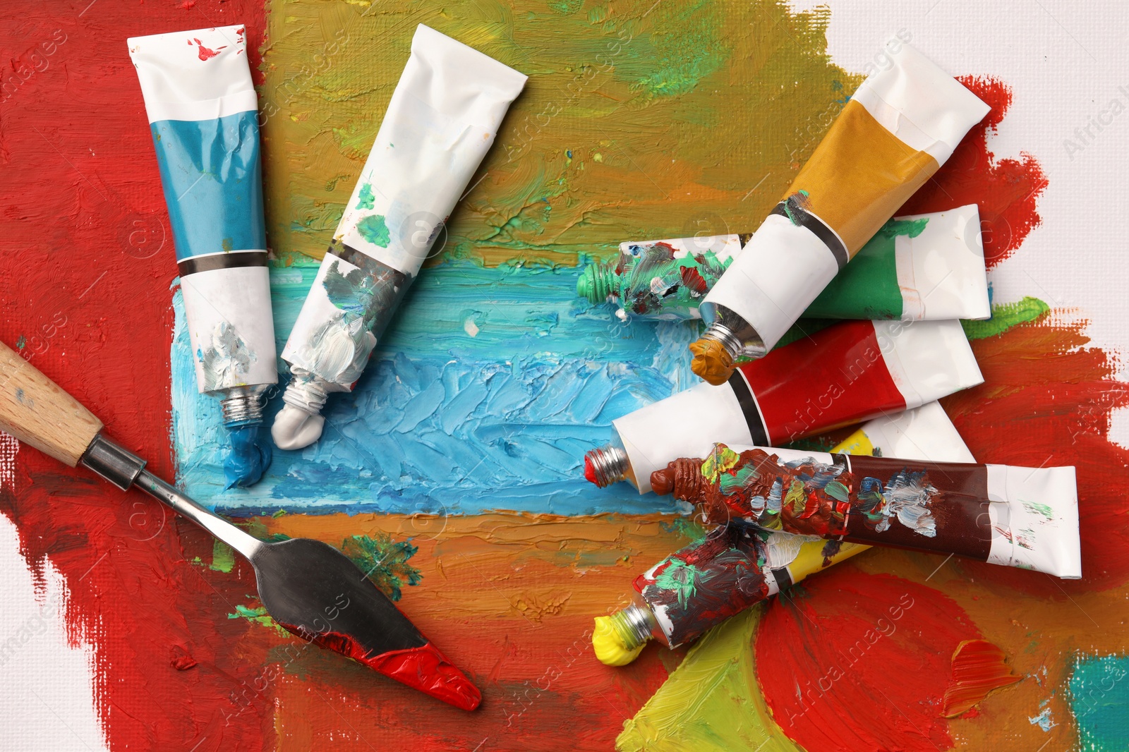 Photo of Tubes of colorful oil paints and spatula on canvas with abstract painting, flat lay