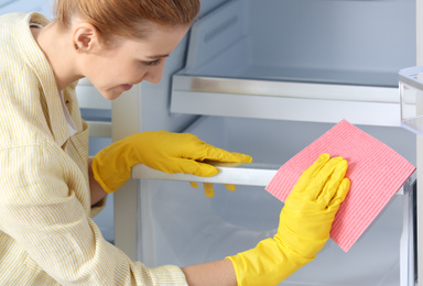 Woman in rubber gloves cleaning empty refrigerator with rag at home