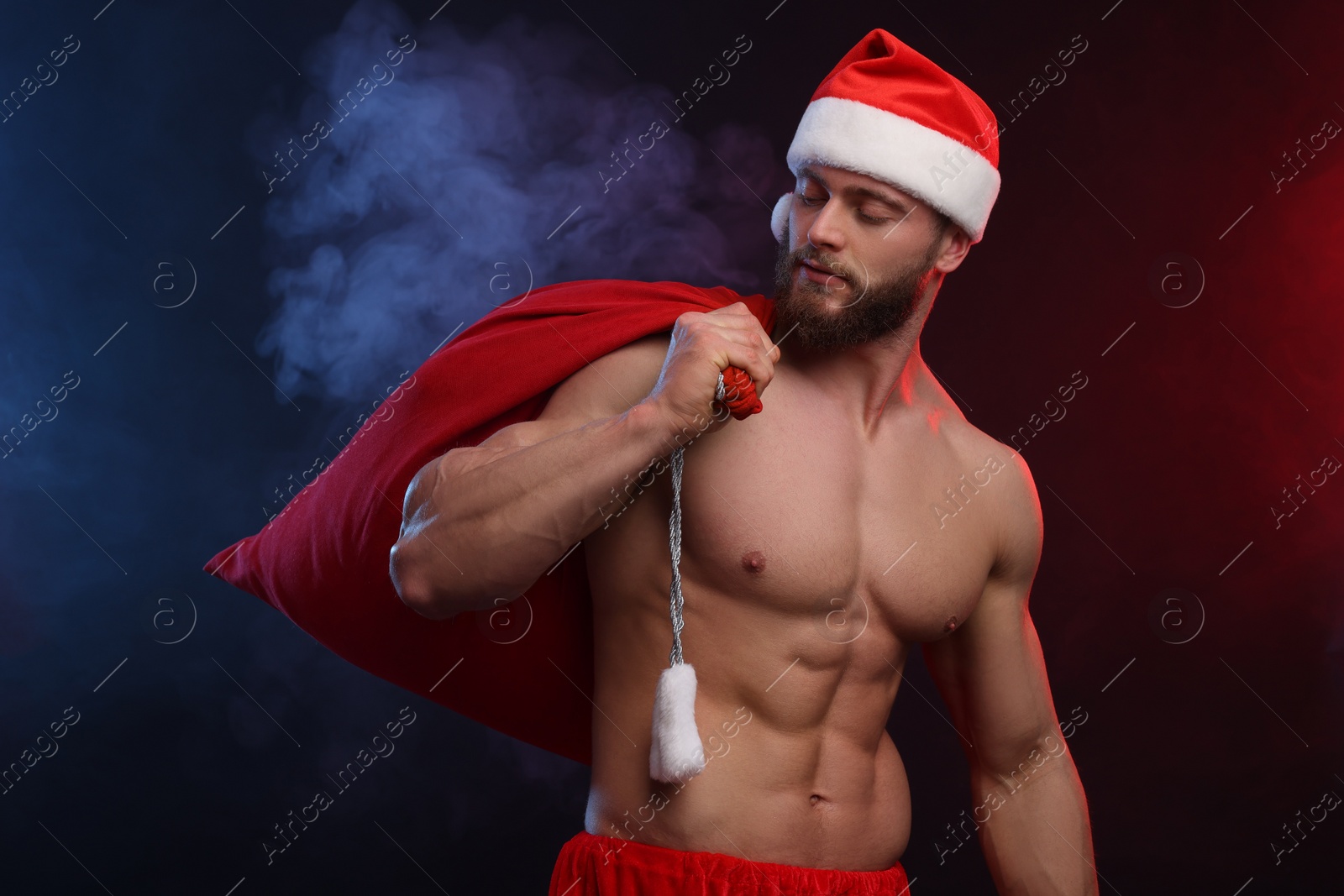 Photo of Muscular young man in Santa hat holding bag with presents on color background