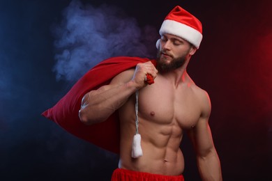 Muscular young man in Santa hat holding bag with presents on color background