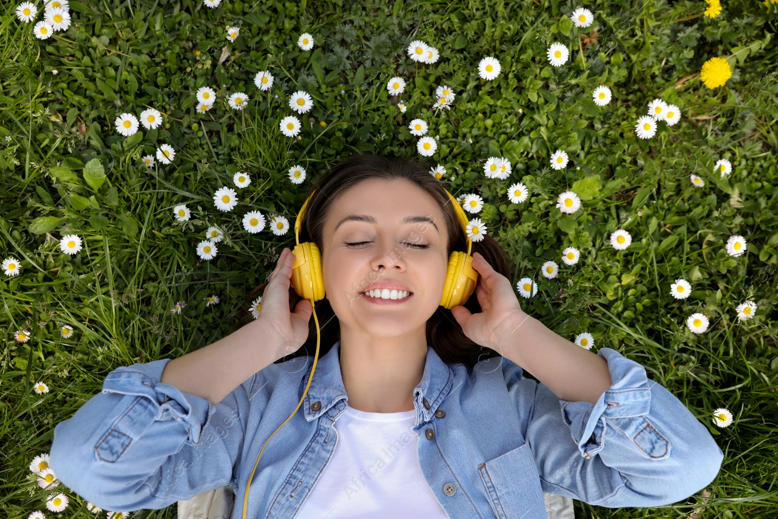 Photo of Happy woman listening to audiobook while lying on grass among blooming daisies, top view