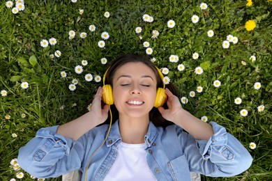 Happy woman listening to audiobook while lying on grass among blooming daisies, top view