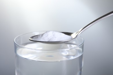 Photo of Spoon with baking soda over glass of water on light grey background, closeup