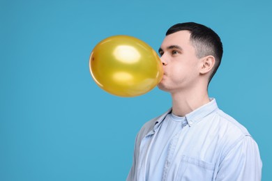 Photo of Young man inflating golden balloon on light blue background. Space for text