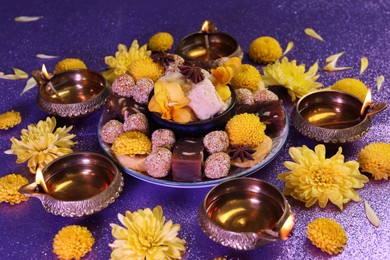 Diwali celebration. Diya lamps, tasty Indian sweets and chrysanthemum flowers on shiny violet table