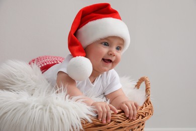 Cute baby wearing Santa hat in wicker basket on light grey background. Christmas celebration