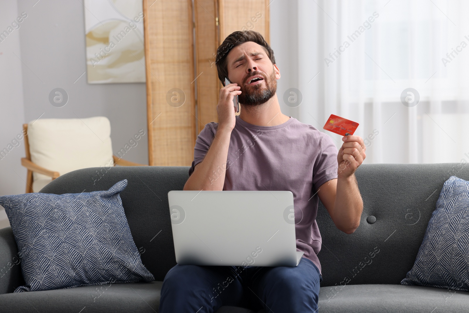 Photo of Emotional man with credit card and laptop talking on phone at home. Be careful - fraud
