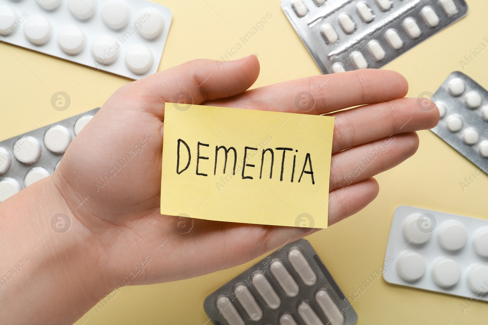 Photo of Woman holding paper note with word Dementia over pills on beige background, top view