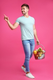 Young man with shopping basket full of products on pink background