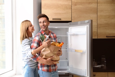 Photo of Happy couple with paper bag full of products near refrigerator in kitchen
