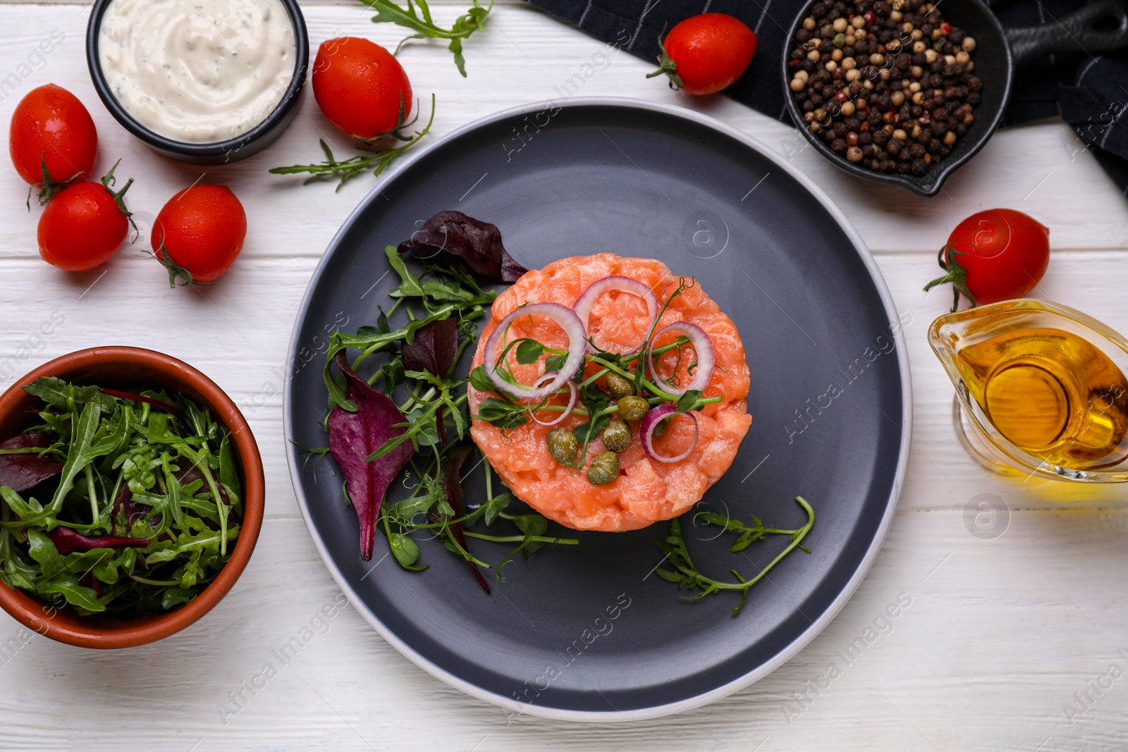 Photo of Tasty salmon tartare with capers, arugula, red onion, basil and microgreens on white wooden table, flat lay