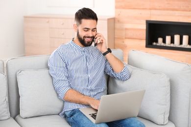 Handsome young man talking on phone while working with laptop on sofa at home