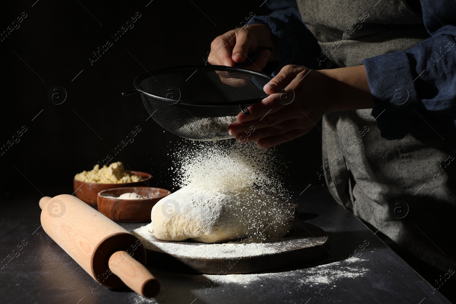 Photo of Woman sprinkling flour over dough at black table, closeup