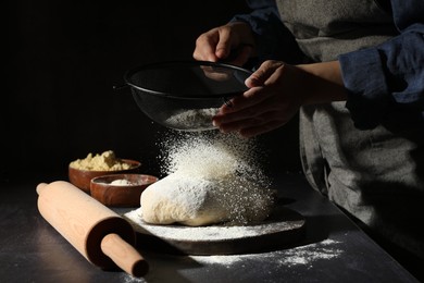 Photo of Woman sprinkling flour over dough at black table, closeup