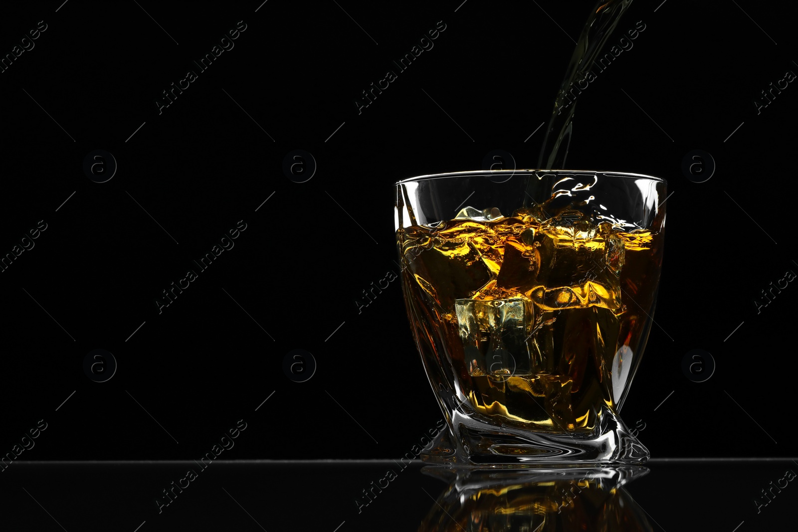 Photo of Pouring whiskey into glass with ice cubes at table against black background, space for text