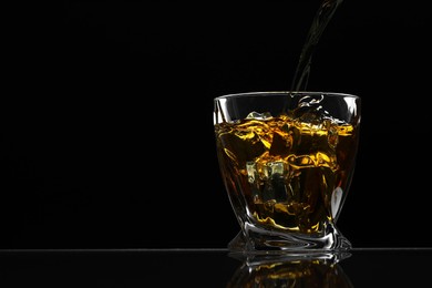 Pouring whiskey into glass with ice cubes at table against black background, space for text