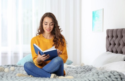 Photo of Young beautiful woman in warm sweater reading book on bed at home