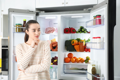 Thoughtful young woman near open refrigerator in kitchen