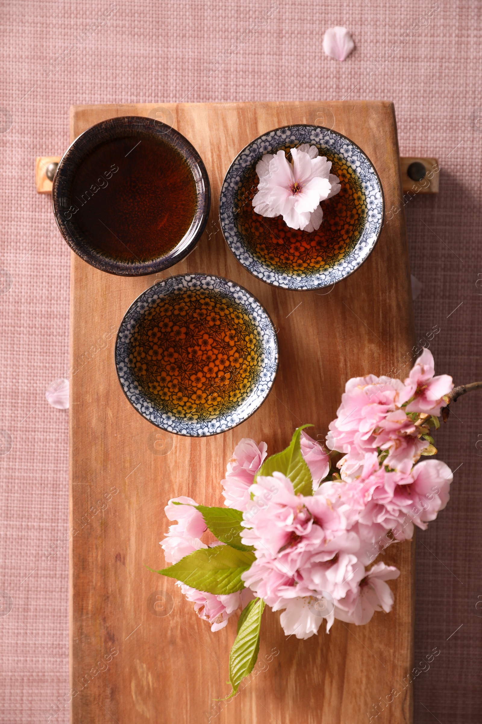 Photo of Wooden tray with cups and sakura flowers on pink fabric, flat lay. Traditional tea ceremony