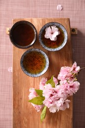 Wooden tray with cups and sakura flowers on pink fabric, flat lay. Traditional tea ceremony