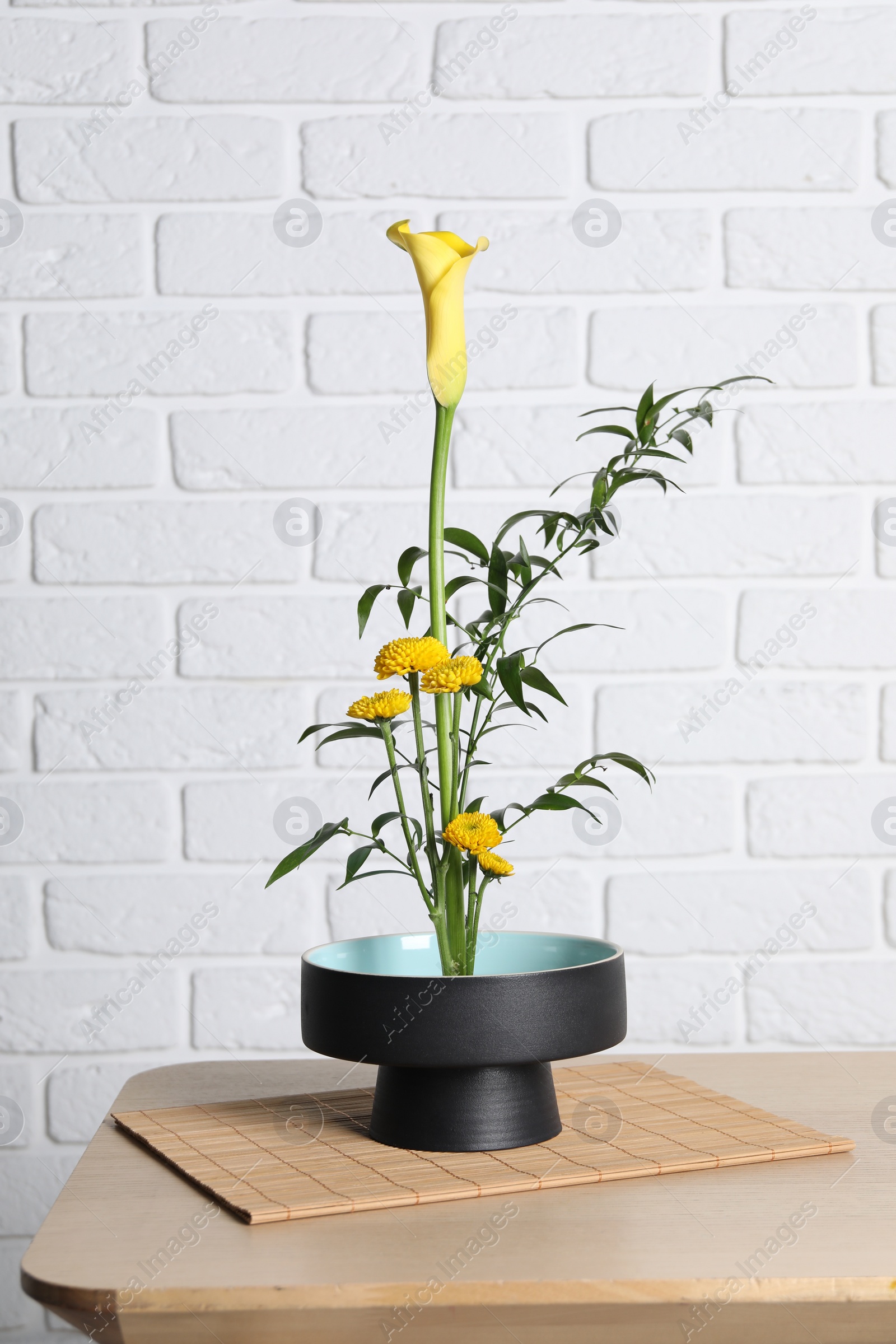 Photo of Beautiful ikebana for stylish house decor. Floral composition with fresh calla, chrysanthemum flowers and branches on wooden table near white brick wall