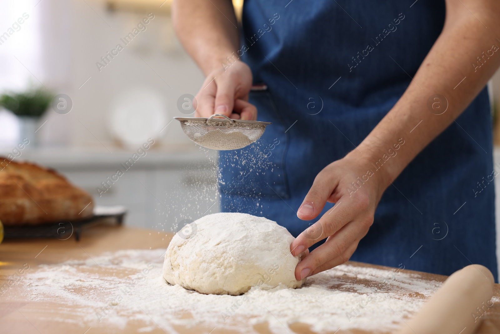 Photo of Making bread. Man sprinkling flour onto dough at wooden table in kitchen, closeup