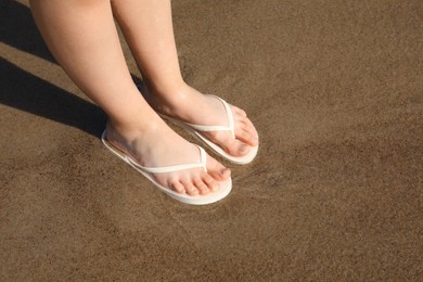 Woman in stylish white flip flops standing on wet sand, closeup