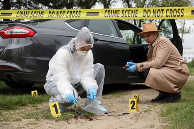 Photo of Investigator and criminologist working with evidences at crime scene near car outdoors