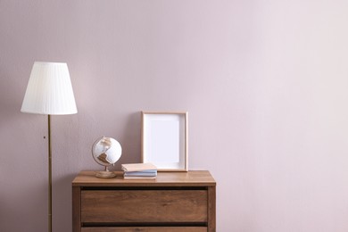 Photo of Wooden chest of drawers with globe, books and empty frame near beige wall in room, space for text. Interior design