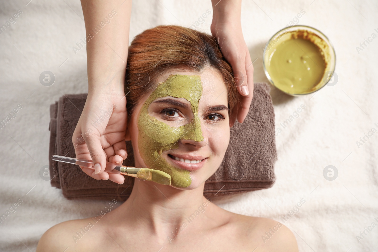 Photo of Cosmetologist applying mask onto woman's face in spa salon, top view