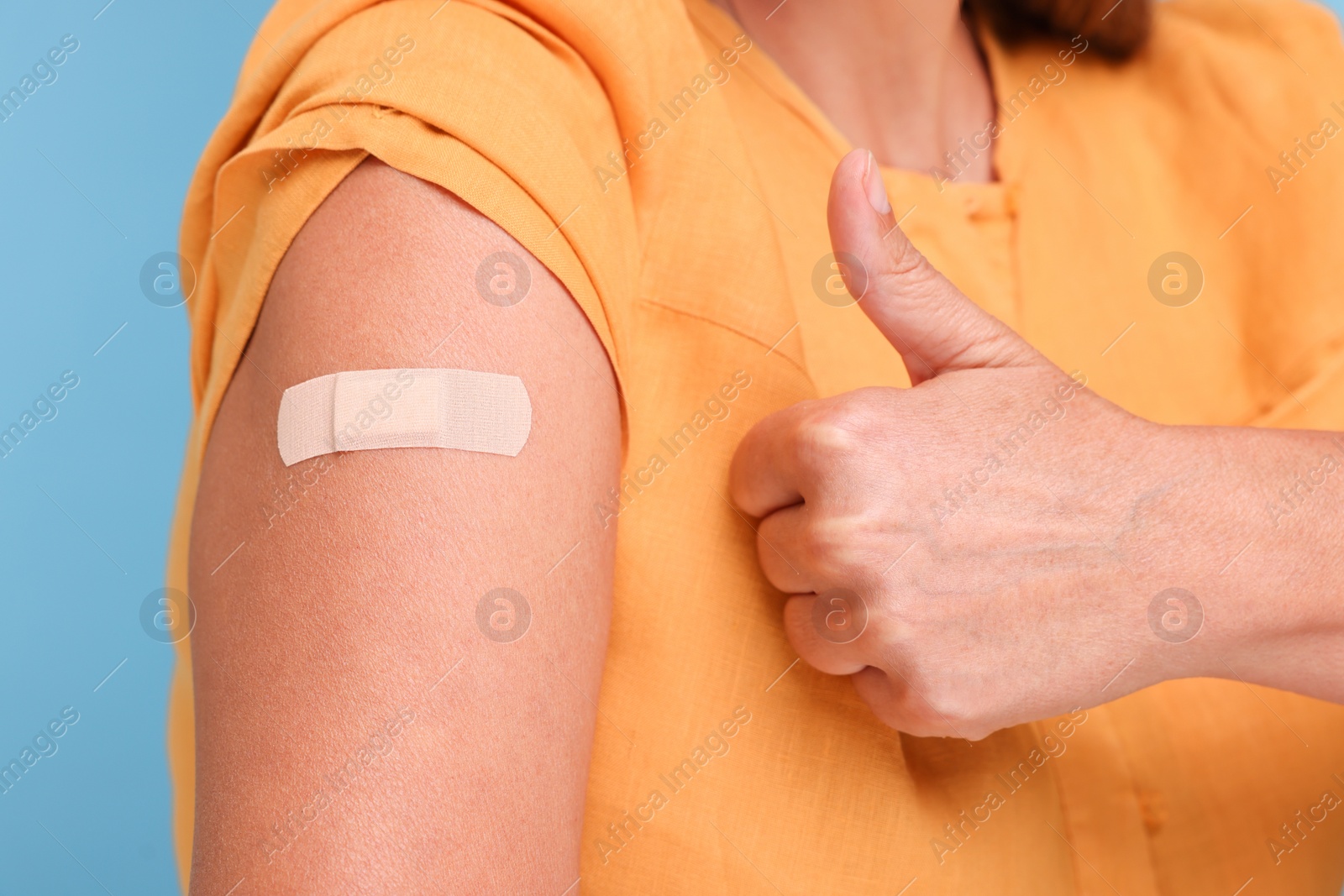 Photo of Woman with adhesive bandage on her arm after vaccination showing thumb up against light blue background, closeup