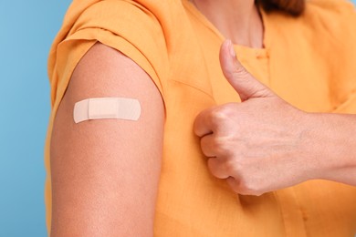 Photo of Woman with adhesive bandage on her arm after vaccination showing thumb up against light blue background, closeup