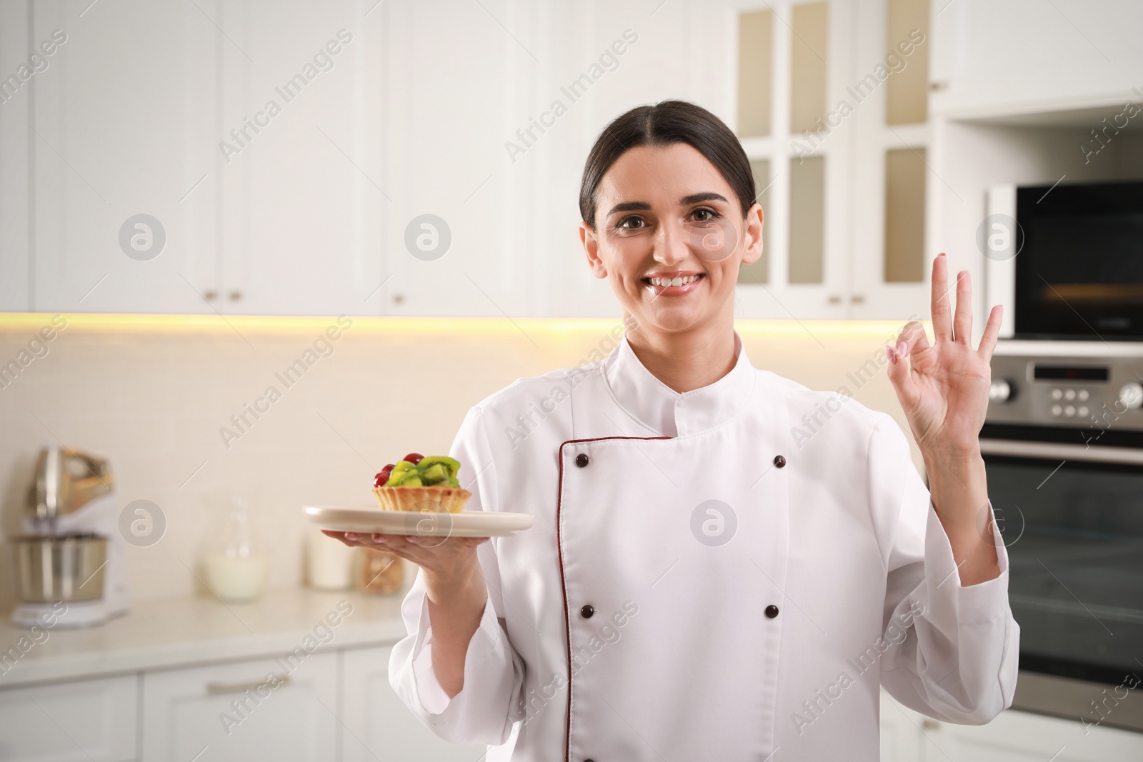 Photo of Happy professional confectioner with tartlet showing okay gesture in kitchen