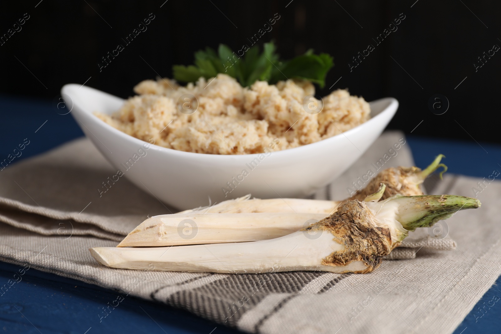 Photo of Bowl of tasty prepared horseradish and roots on blue wooden table, closeup