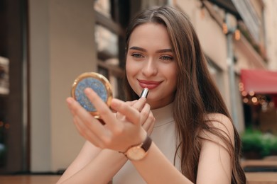 Photo of Beautiful young woman with cosmetic pocket mirror applying lipstick outdoors