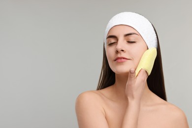 Young woman with headband washing her face using sponge on light grey background, space for text