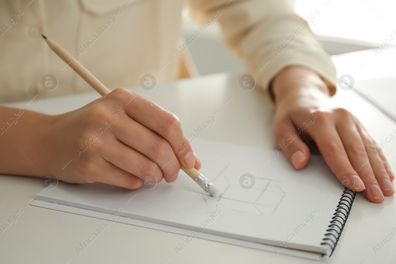 Photo of Woman correcting picture in notepad with pencil eraser at white table, closeup