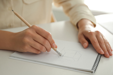 Photo of Woman correcting picture in notepad with pencil eraser at white table, closeup