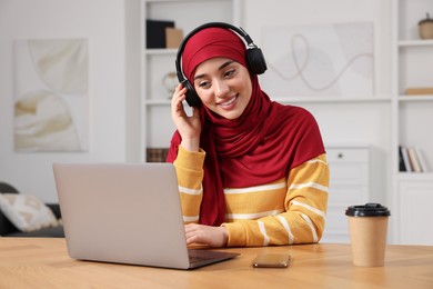 Photo of Muslim woman in hijab with headphones using laptop at wooden table in room