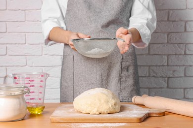 Photo of Woman sprinkling flour over dough at table near white brick wall, closeup