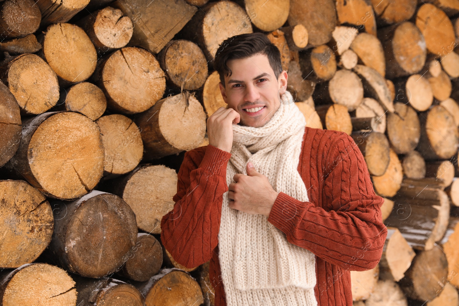 Photo of Handsome man wearing warm sweater and scarf near stack of firewood. Winter season