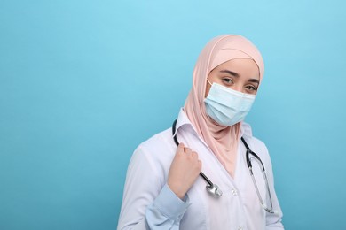 Photo of Muslim woman wearing hijab, medical uniform and protective mask on light blue background, space for text