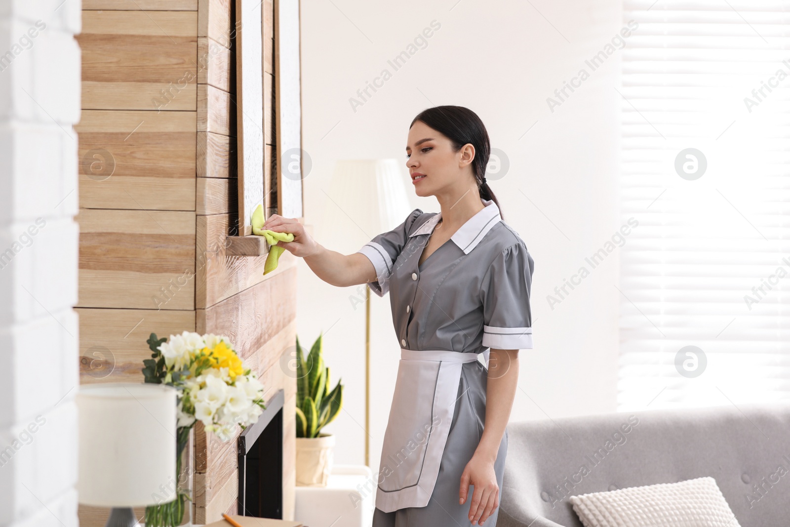 Photo of Young chambermaid wiping dust from furniture in hotel room