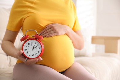 Photo of Young pregnant woman holding alarm clock near her belly at home, closeup. Time to give birth