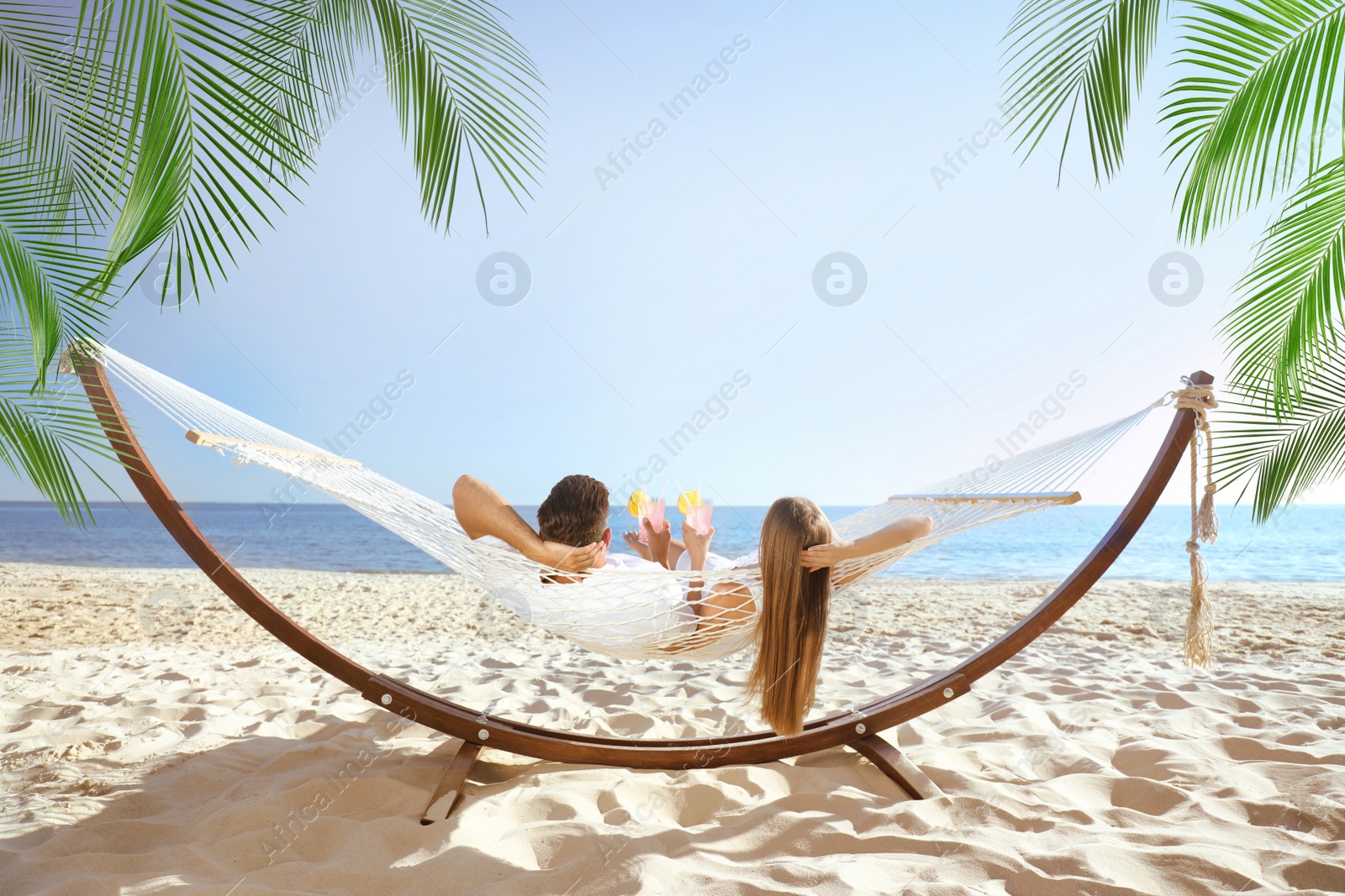 Image of Couple with refreshing cocktails relaxing in hammock under green palm leaves on beach