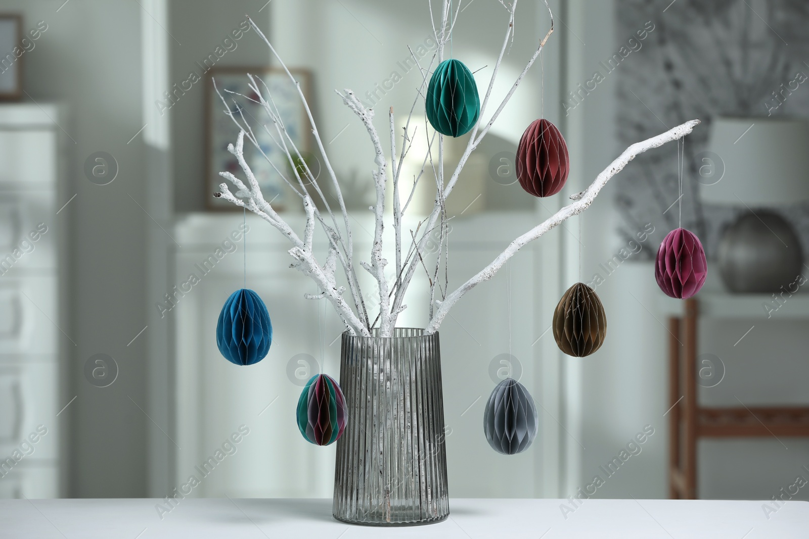 Photo of Branches with paper eggs in vase on white table at home. Beautiful Easter decor